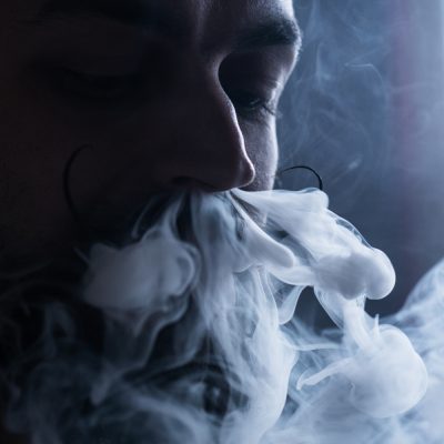 Young Man with Beard and Mustages Vaping an Electronic Cigarette. Vaper Hipster Smoke Vaporizer and Exhals Smoke Flow from Nose.