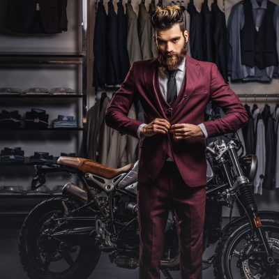 Elegantly dressed bearded man buttons jacket near standing retro sports motorbike at men's clothing store.